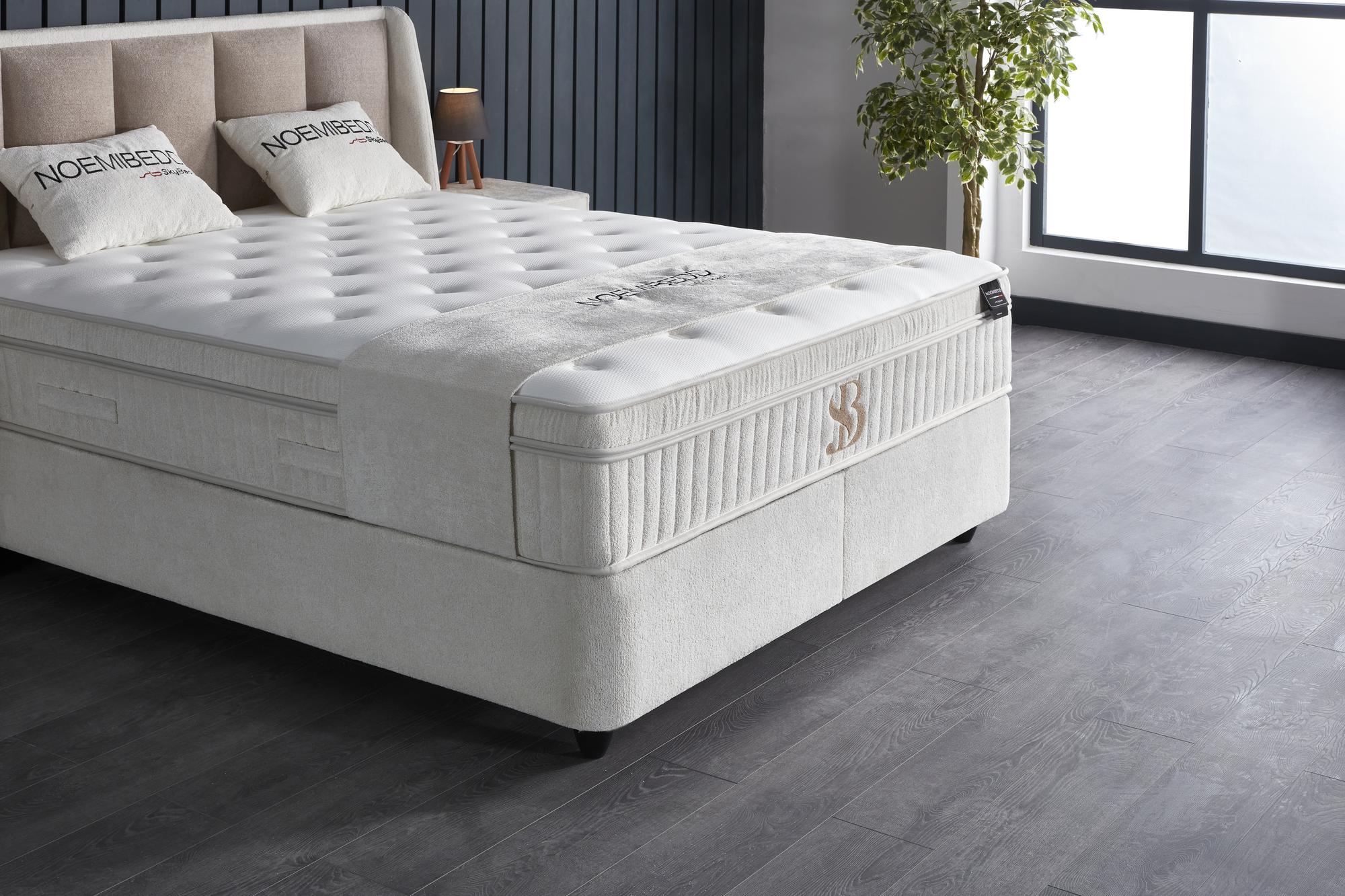Boxspringbett NoemiBedd mit integrierter Matratze und Stauraum von SkyHome24.de, ideal für günstigen, komfortablen Schlaf, stilvoll präsentiert mit einem Kissen und Pflanze.