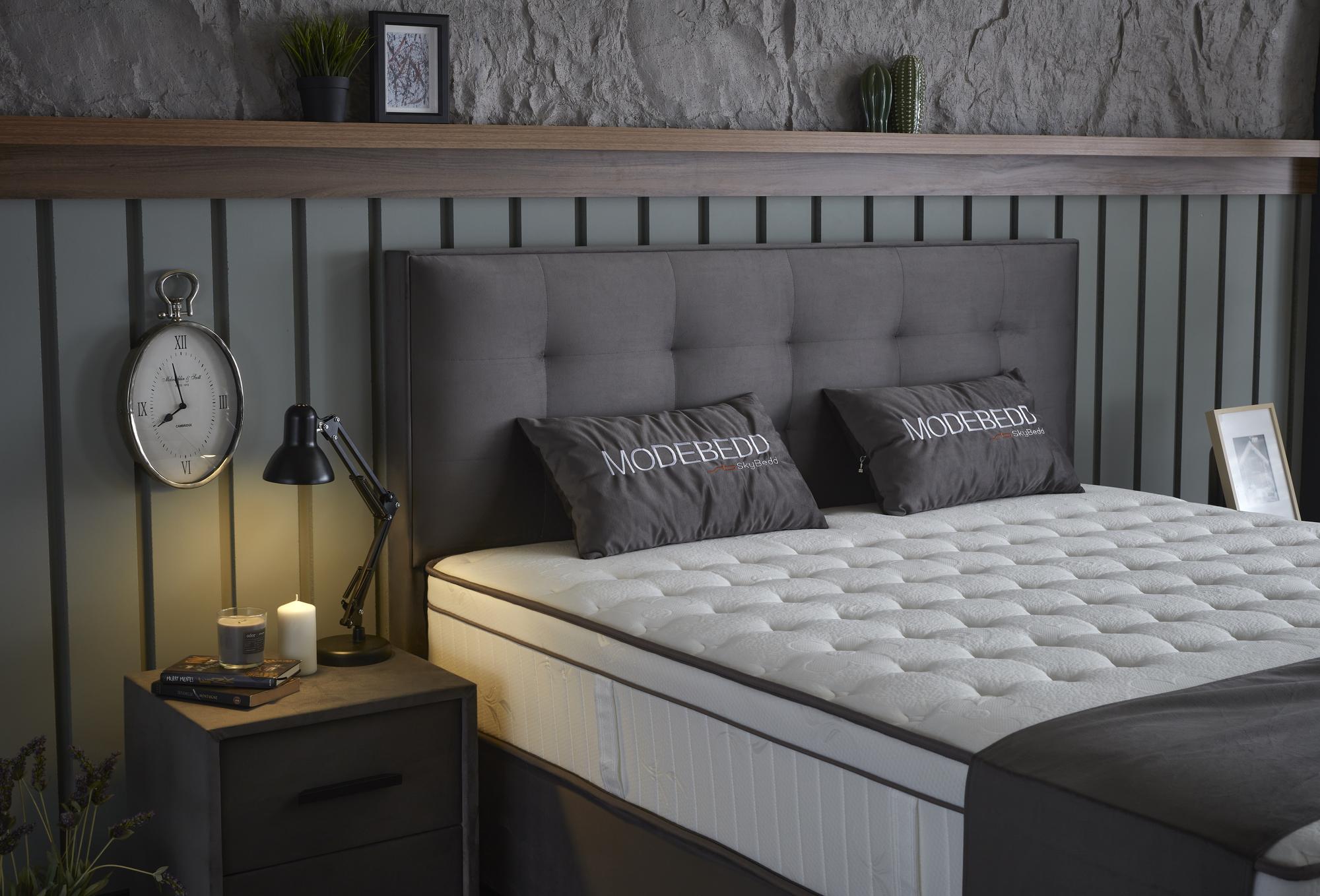 Boxspringbett 'ModeBedd' SkyBedd in Grau/Anthrazit mit ergonomischer Matratze und integriertem Stauraum, präsentiert in einem stilvollen Innenraum mit Uhr und Bild.