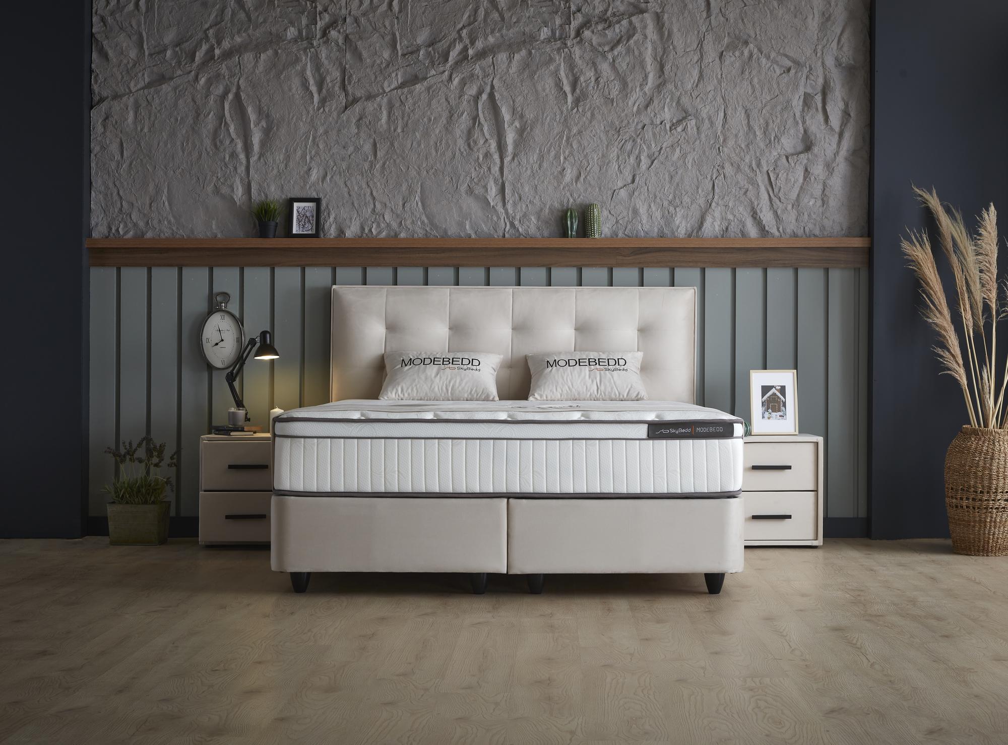 Boxspringbett 'ModeBedd' SkyBedd mit ergonomischer Matratze und integriertem Stauraum, umgeben von stilvollen Dekorationen wie einer Pflanze, einem Kissen und einer Uhr.