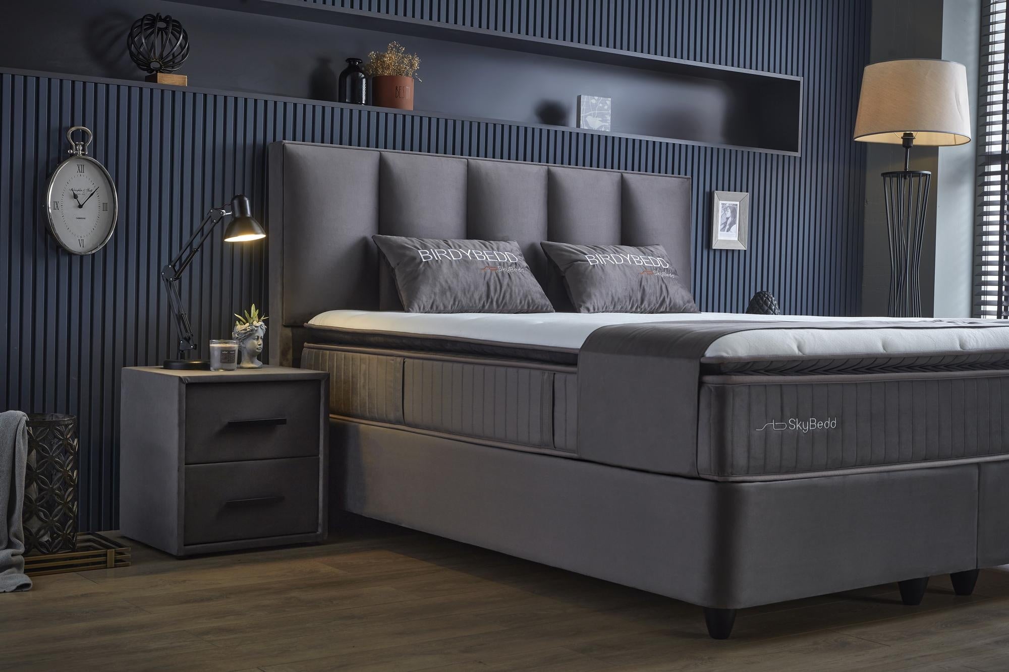 Boxspringbett 'BirdyBedd' SkyBedd in Grau mit Matratze und integriertem Stauraum, umgeben von einer eleganten Inneneinrichtung mit Uhr, Lampe und Bilderrahmen.