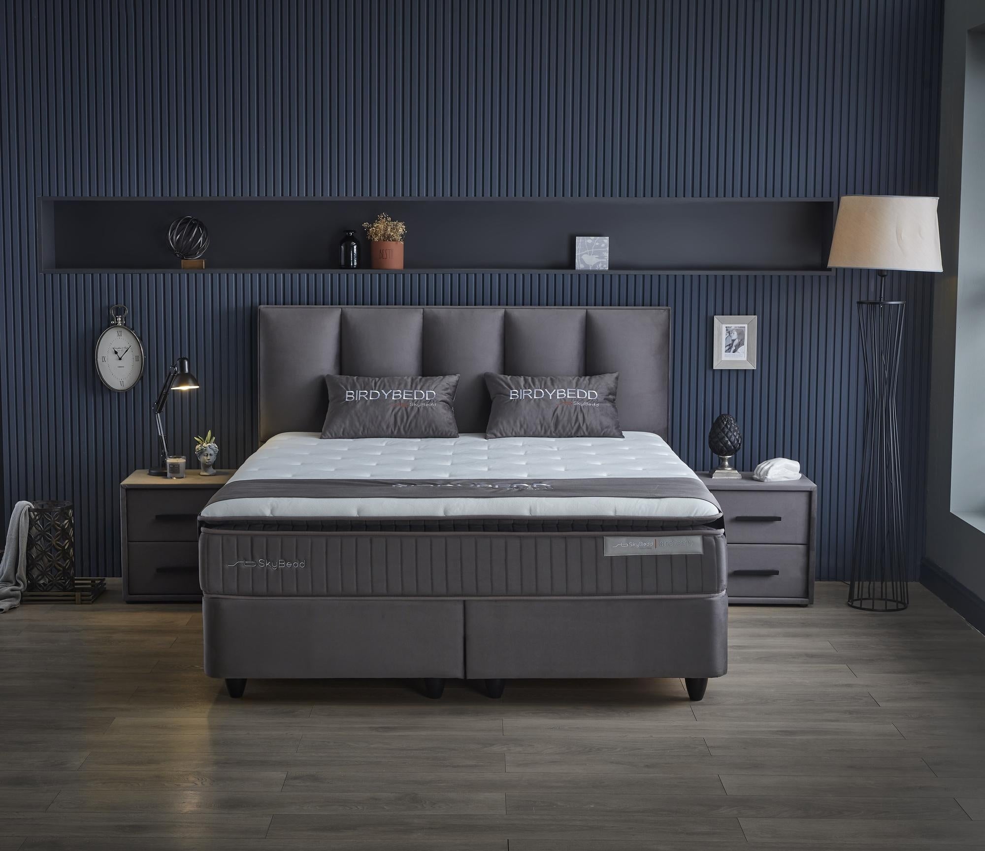 Boxspringbett 'BirdyBedd' SkyBedd mit gepolsterter Matratze und integriertem Stauraum, präsentiert in einem stilvoll eingerichteten Raum mit Wanduhr und Bilderrahmen.