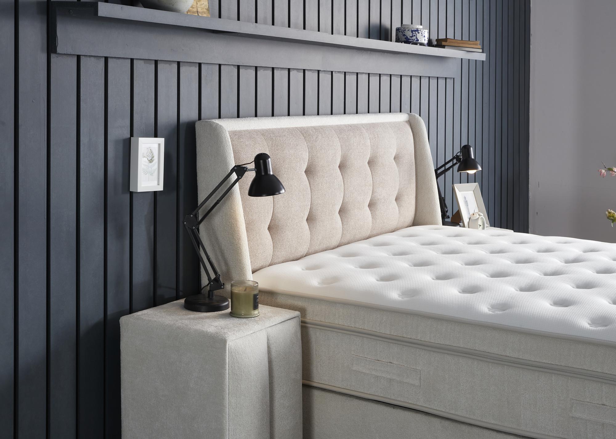 Boxspringbett 'PerlaBedd' von SkyHome24.de mit integrierter Matratze und Stauraum, neben einem Bett mit schwarzer Lampe und einem Bild im Hintergrund.