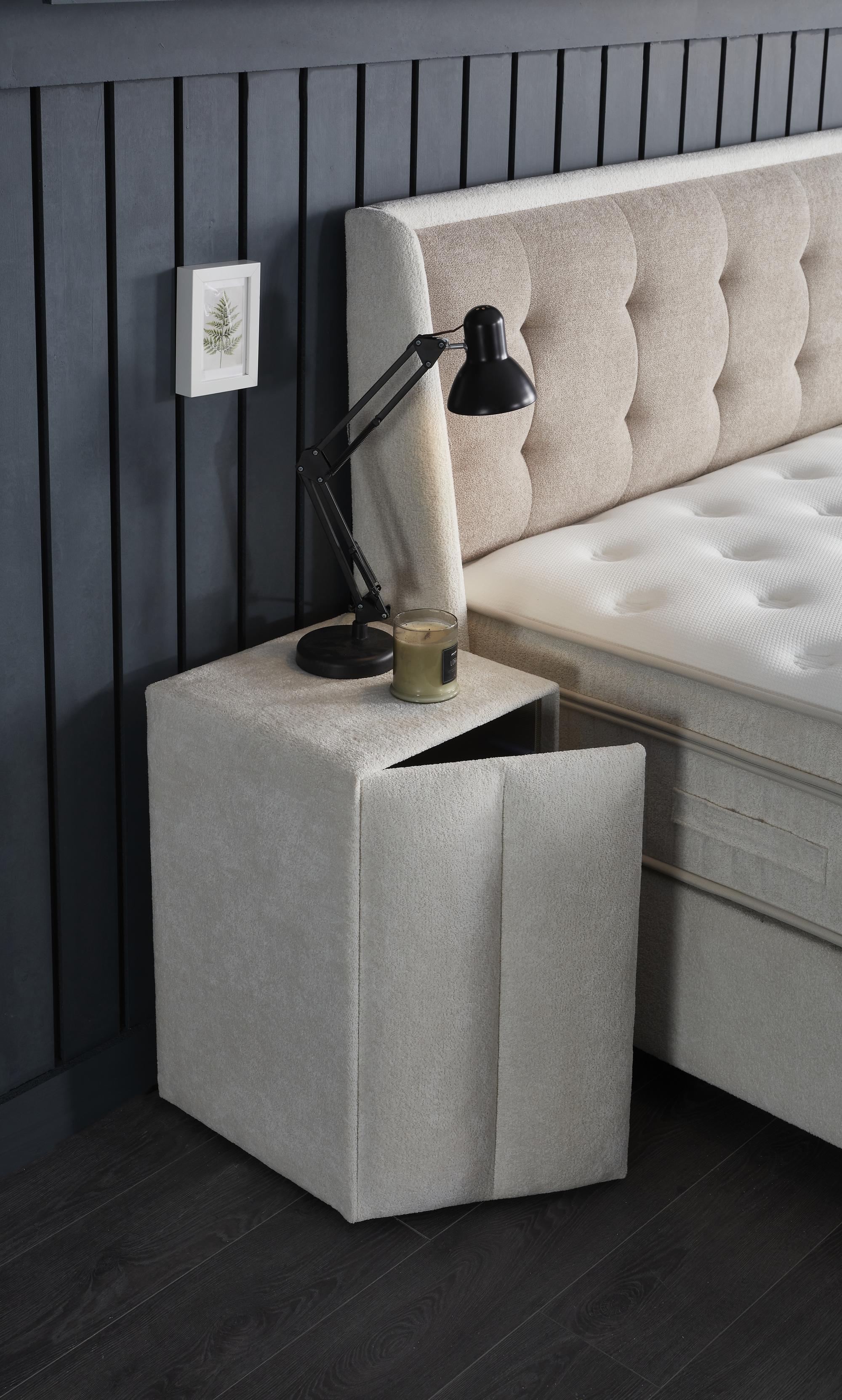 Boxspringbett 'PerlaBedd' von SkyHome24.de in Beige mit Matratze und integriertem Stauraum, neben einer Lampe und Kerze platziert.